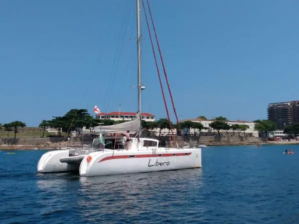 Catamarã de 47 pés ancorado em águas calmas, visto de perfil, com mastro alto e vela recolhida, orla urbana ao fundo
