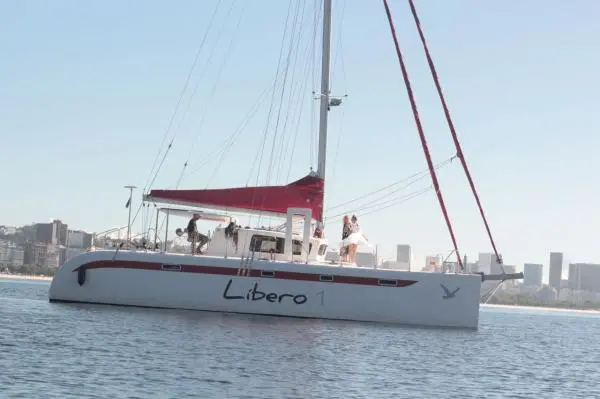 Catamarã Praia 47 a vela na orla do Rio de Janeiro, com pessoas a bordo e prédios da cidade ao fundo