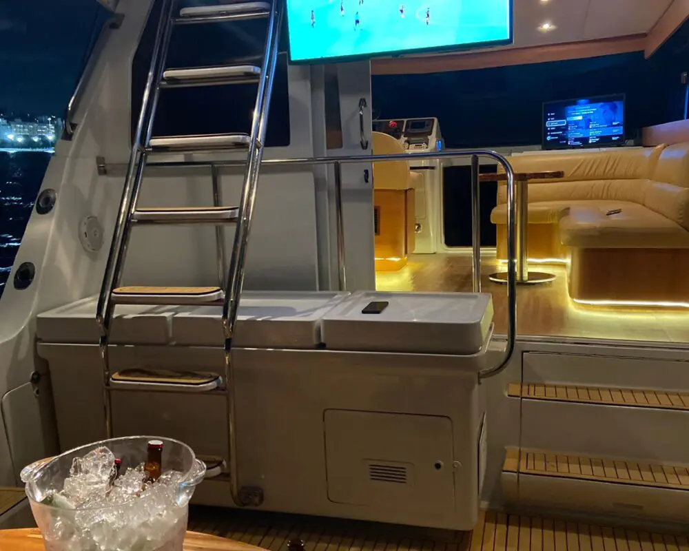 Área externa da Lancha Rio Star 50 à noite, com TV ligada exibindo futebol, mesa com bebidas geladas e ambiente iluminado.