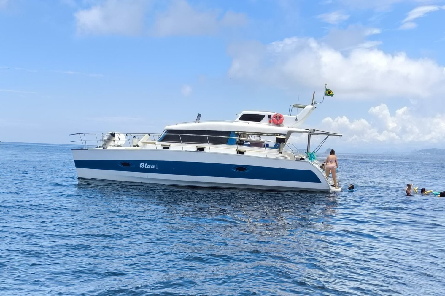 Catamarã Powercat 40 fundeado em mar aberto, com escada de popa em uso e pessoas nadando ao lado do casco