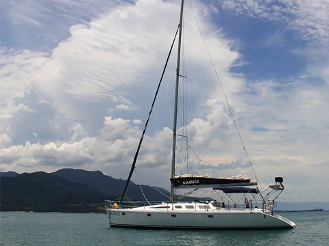 Veleiro One Off 47 fundeado em mar aberto, com mastro, bimini armado e costa montanhosa ao fundo