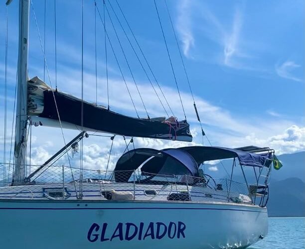 Casco branco do veleiro MJ 38 fundeado em água esverdeada, com bimini azul e mastro sob céu limpo