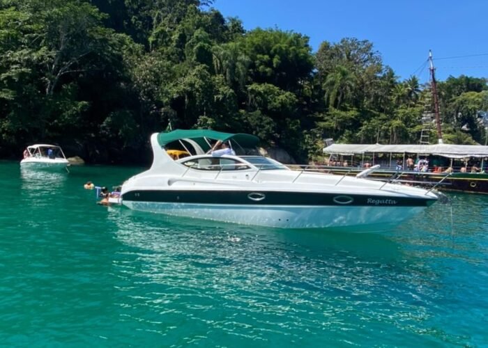 lancha Real 29 ancorada em água verde-esmeralda junto a costão com mata, com banhistas e escuna ao lado