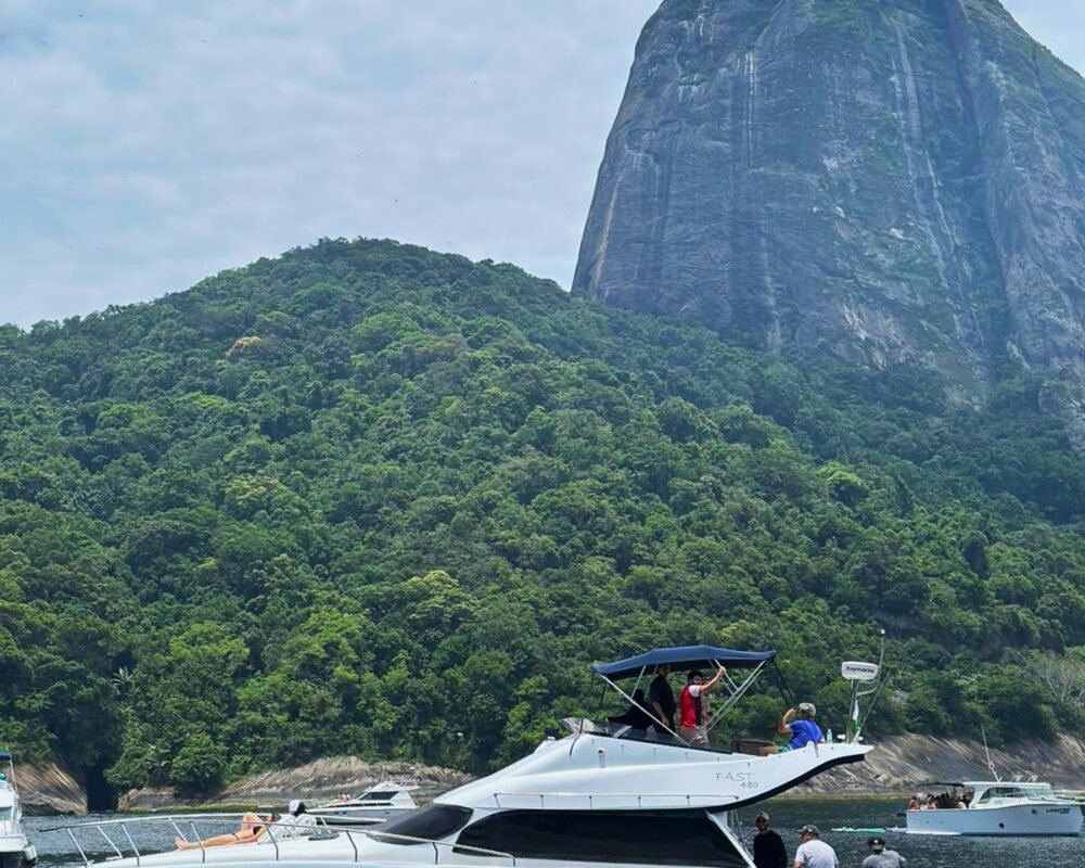Lancha Fast Senna 48 navegando próximo ao Pão de Açúcar, no Rio de Janeiro, com vegetação exuberante ao fundo. Algumas pessoas estão aproveitando o passeio na lancha.