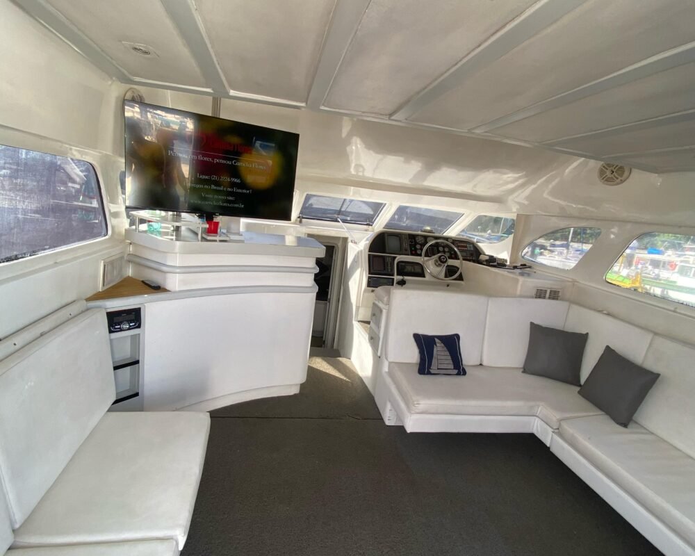 Salão interno da lancha com sofás brancos, TV e espaço amplo para socializar. Perfeito para eventos e confraternizações durante o passeio.