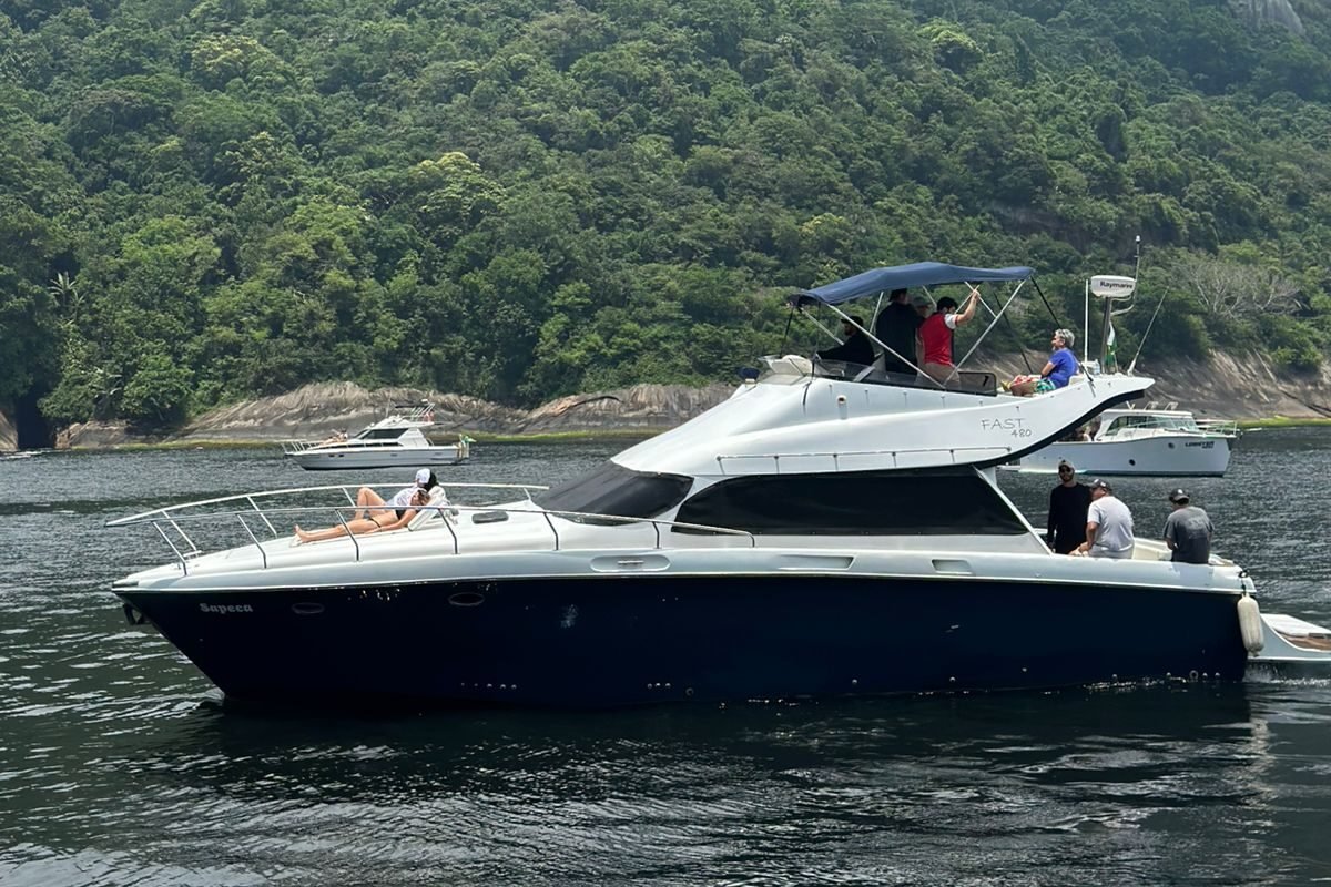 Lancha Fast Senna 48 navegando próximo ao Pão de Açúcar, no Rio de Janeiro, com vegetação exuberante ao fundo. Algumas pessoas estão aproveitando o passeio na lancha.