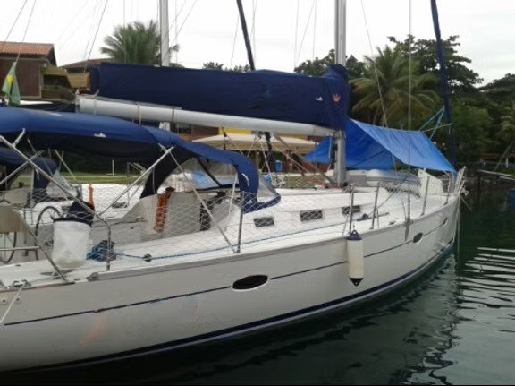 Veleiro de casco branco atracado na marina, com capas de vela e capota azuis, velas recolhidas e vegetação ao fundo.