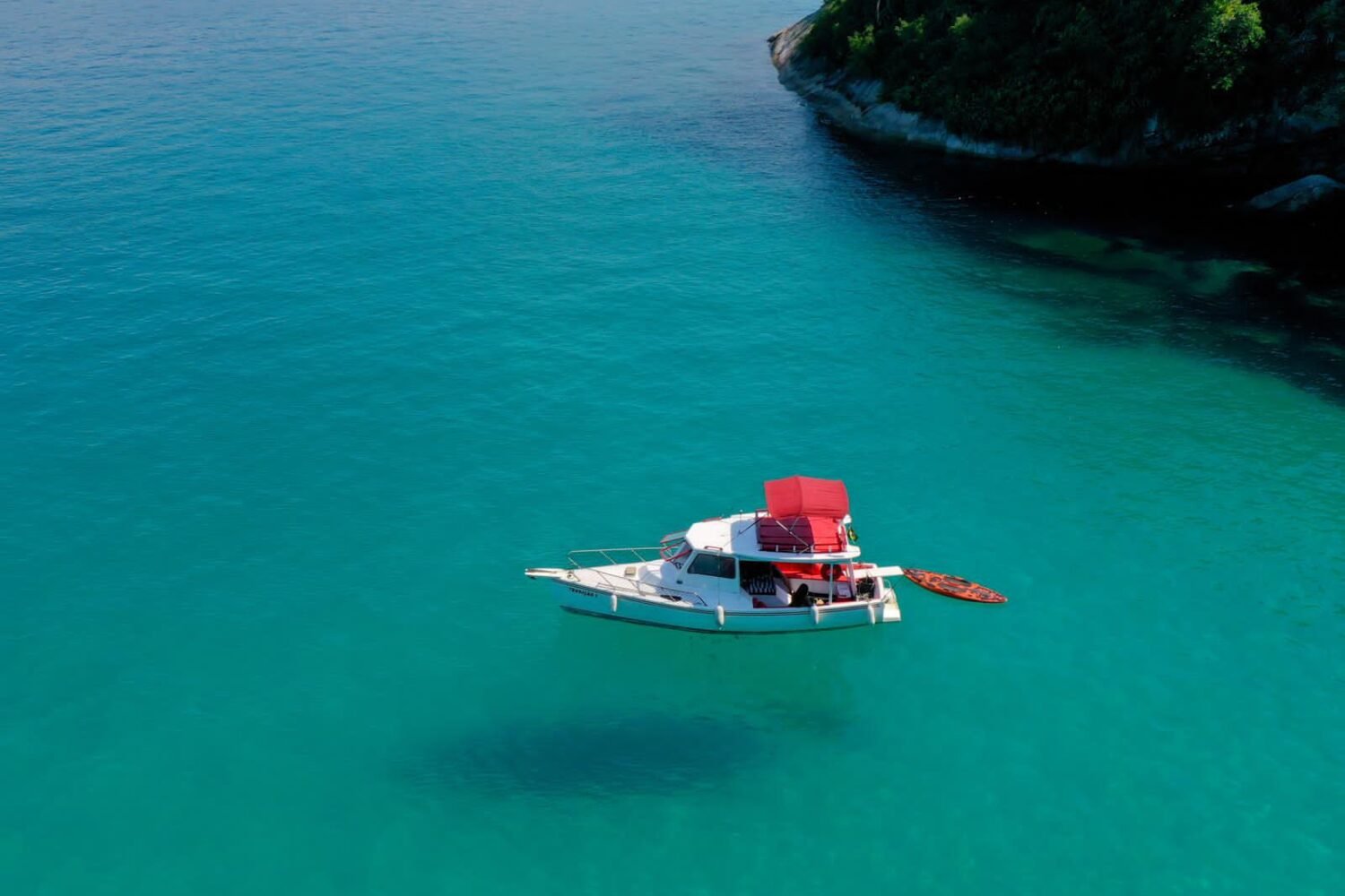 Barco traineira ancorado em águas cristalinas de Paraty, com um stand-up paddle ao lado.