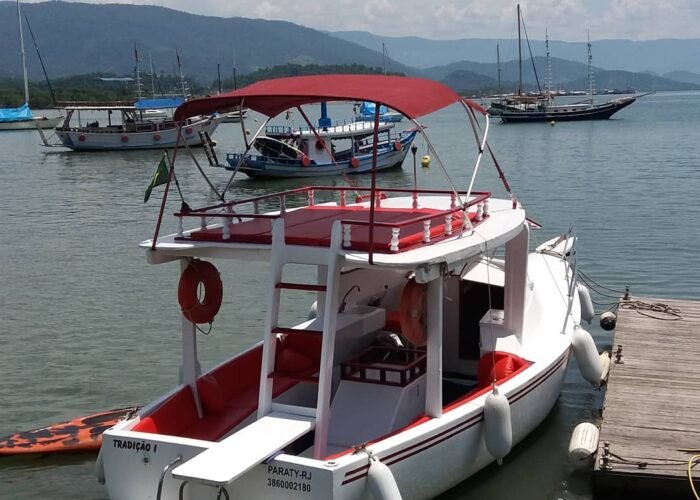 traineira com toldo vermelho e solário superior atracada em píer de Paraty, com escunas ancoradas e montanhas ao fundo