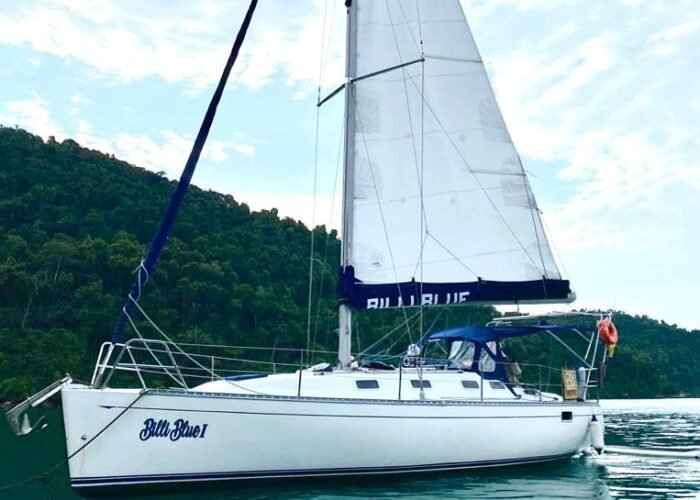 Veleiro B&B 36 de perfil navegando com vela grande e genoa, próximo a morro coberto de mata
