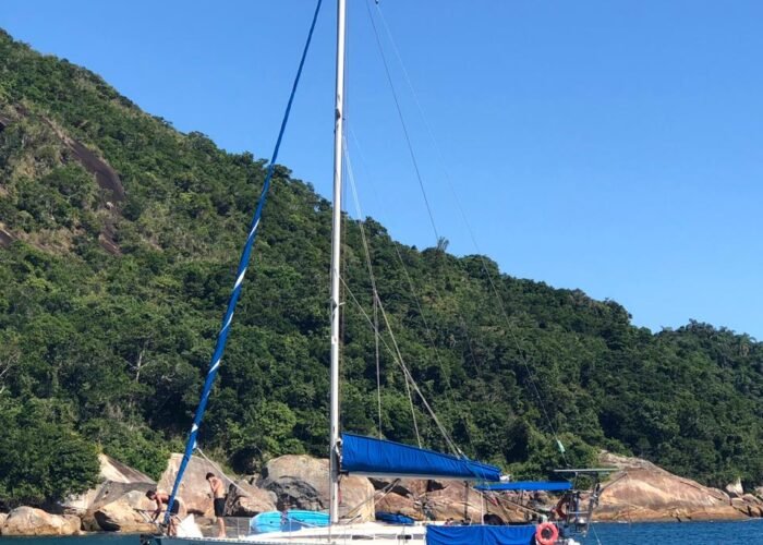 Veleiro Fast 395 branco fundeado em enseada de mata fechada, com capa de vela azul, bimini e bote de apoio ao lado