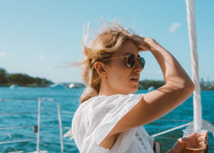 Mulher loira com óculos escuros e roupa leve aproveitando um passeio de lancha sob o sol, com o mar azul ao fundo. Look confortável e estiloso para curtir um dia na embarcação.