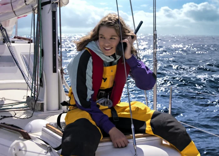 Jessica Watson em um barco à vela, usando roupa de proteção amarela e falando pelo rádio, navegando pelo oceano em um dia ensolarado.