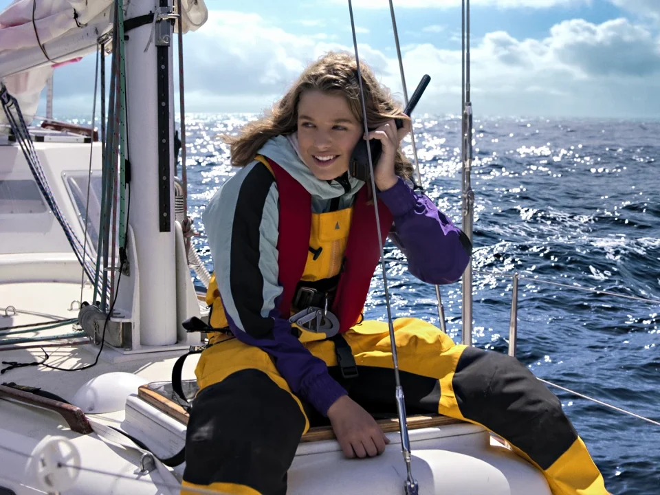 Jessica Watson em um barco à vela, usando roupa de proteção amarela e falando pelo rádio, navegando pelo oceano em um dia ensolarado.