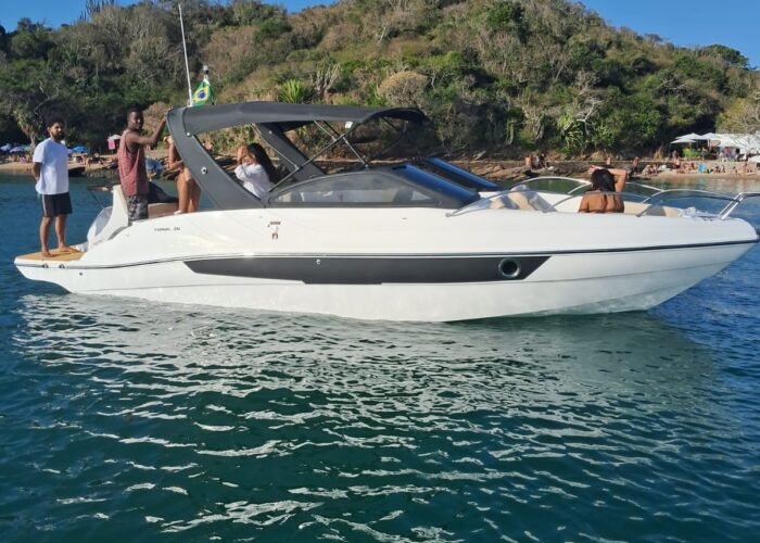 Lancha branca de 26 pés navegando em águas calmas de Búzios, com costão verde ao fundo e praia ao longe