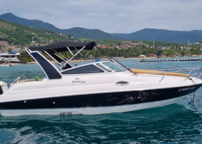 Lancha Focker 255 de perfil, com casco preto e branco, toldo bimini e solário de proa, fundeada perto da praia