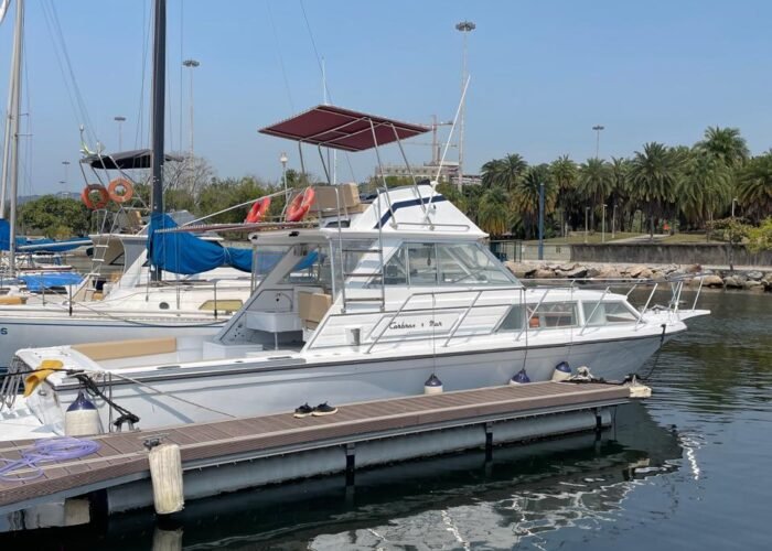 Lancha branca atracada na marina, vista de perfil, com toldo bordô sobre o flybridge e vegetação ao fundo.