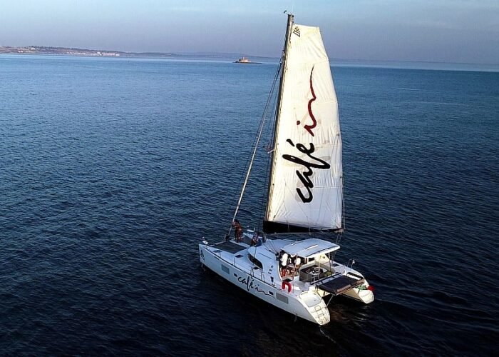 Vista aérea do catamarã Lagoon 380 S2 navegando à vela em águas calmas, com a costa no horizonte