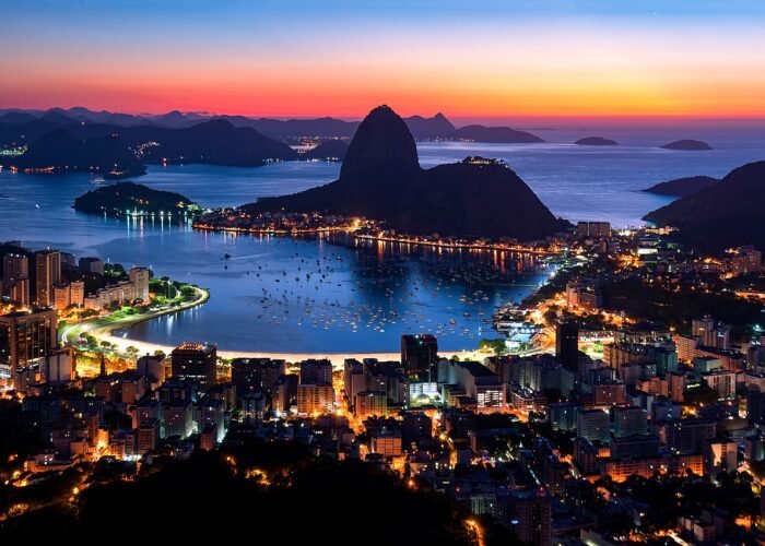 Vista panorâmica do Rio de Janeiro ao anoitecer, destacando o Pão de Açúcar, a Baía de Guanabara com barcos ancorados e a cidade iluminada. O céu exibe tons alaranjados e azulados, criando um cenário deslumbrante.