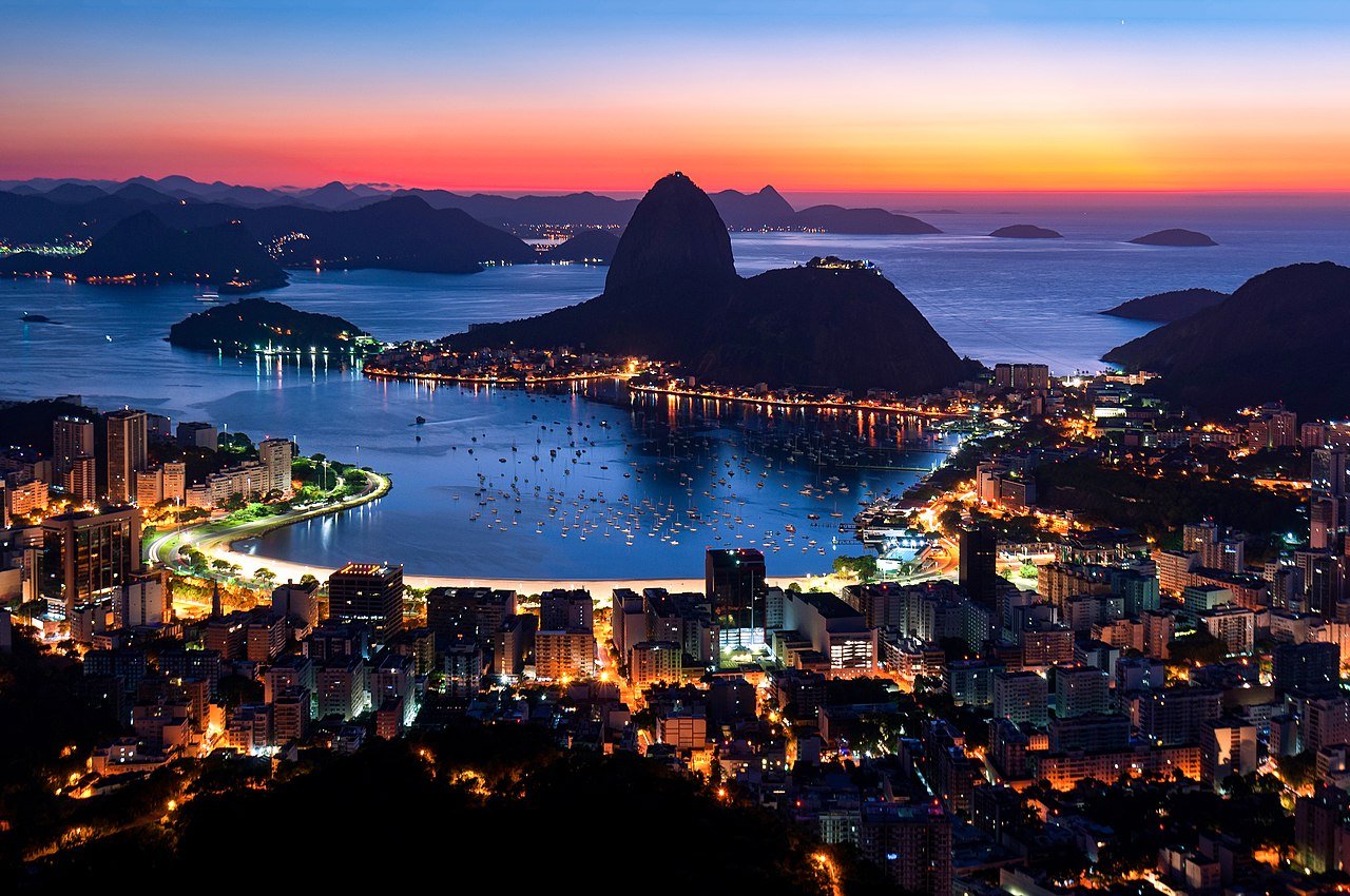 Vista panorâmica do Rio de Janeiro ao anoitecer, destacando o Pão de Açúcar, a Baía de Guanabara com barcos ancorados e a cidade iluminada. O céu exibe tons alaranjados e azulados, criando um cenário deslumbrante.