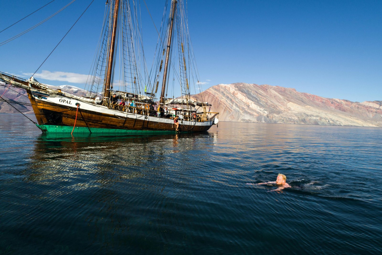 Escuna de madeira ancorada em águas calmas, com turistas a bordo e um nadador aproveitando o mar cristalino. Ao fundo, montanhas formam uma paisagem impressionante.