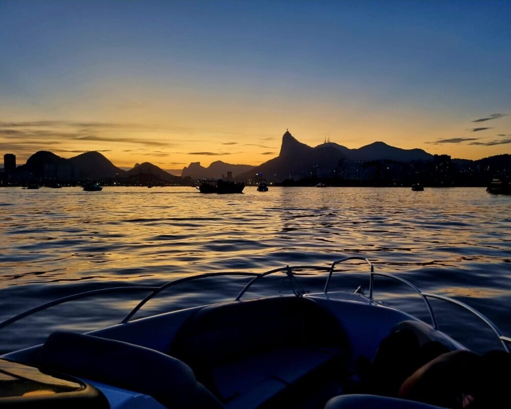 Pôr do sol visto do convés da lancha Coral 26, com reflexos dourados no mar e montanhas ao fundo.