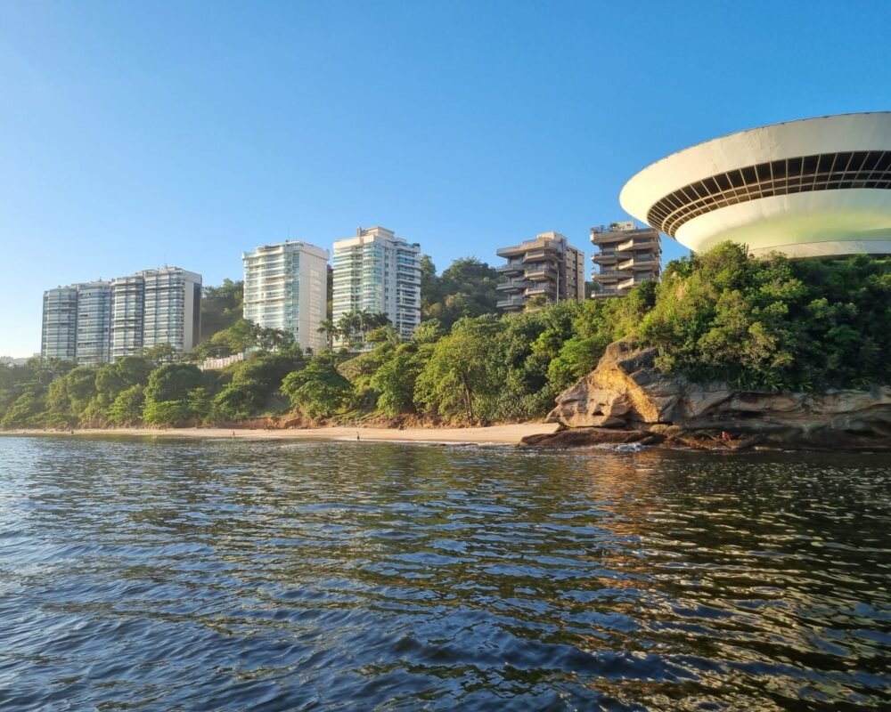 Museu de Arte Contemporânea (MAC) de Niterói visto do mar, rodeado por vegetação e prédios modernos.