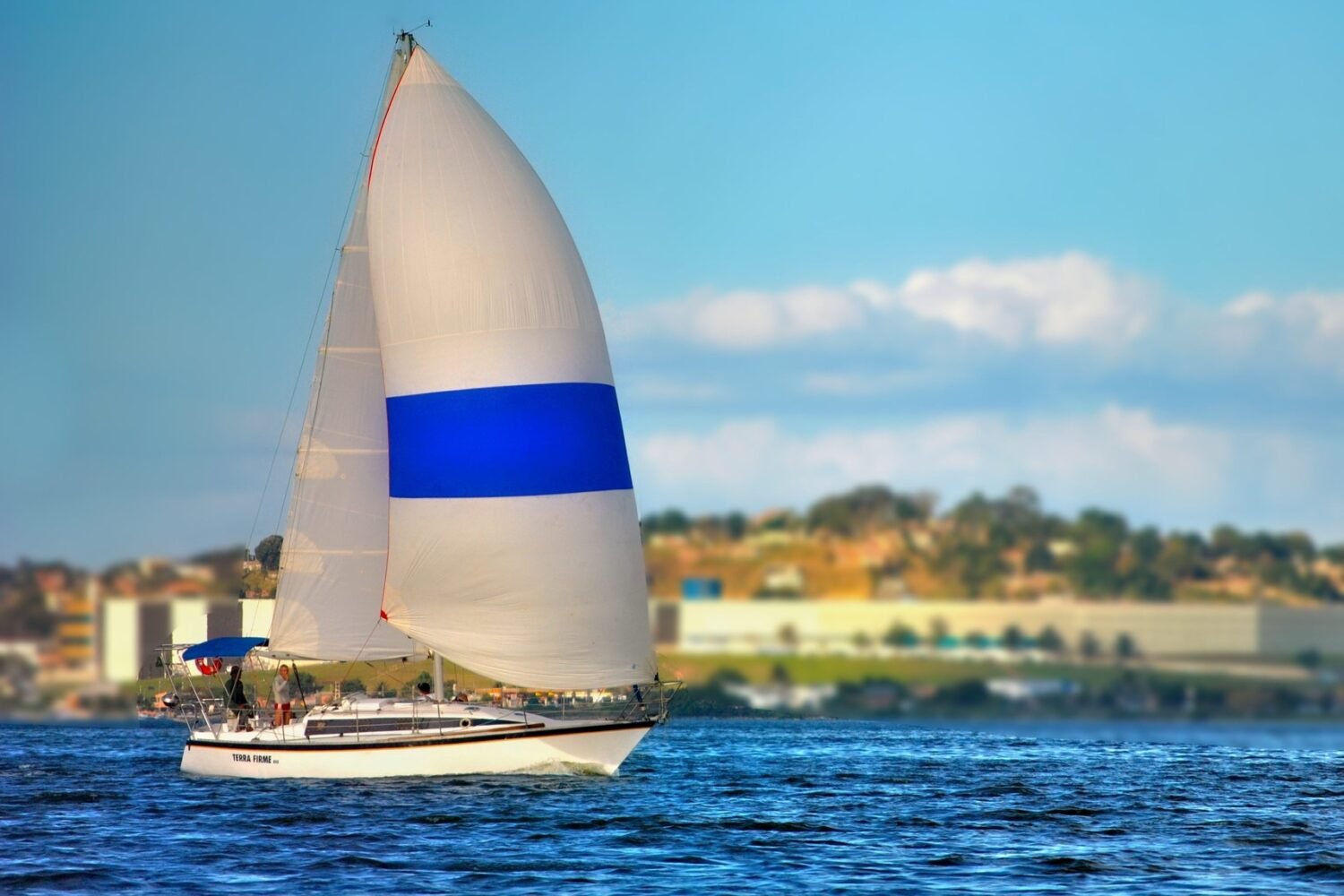 Veleiro Peterson 33 navegando com balão branco e azul aberto em mar azul, com a costa ao fundo