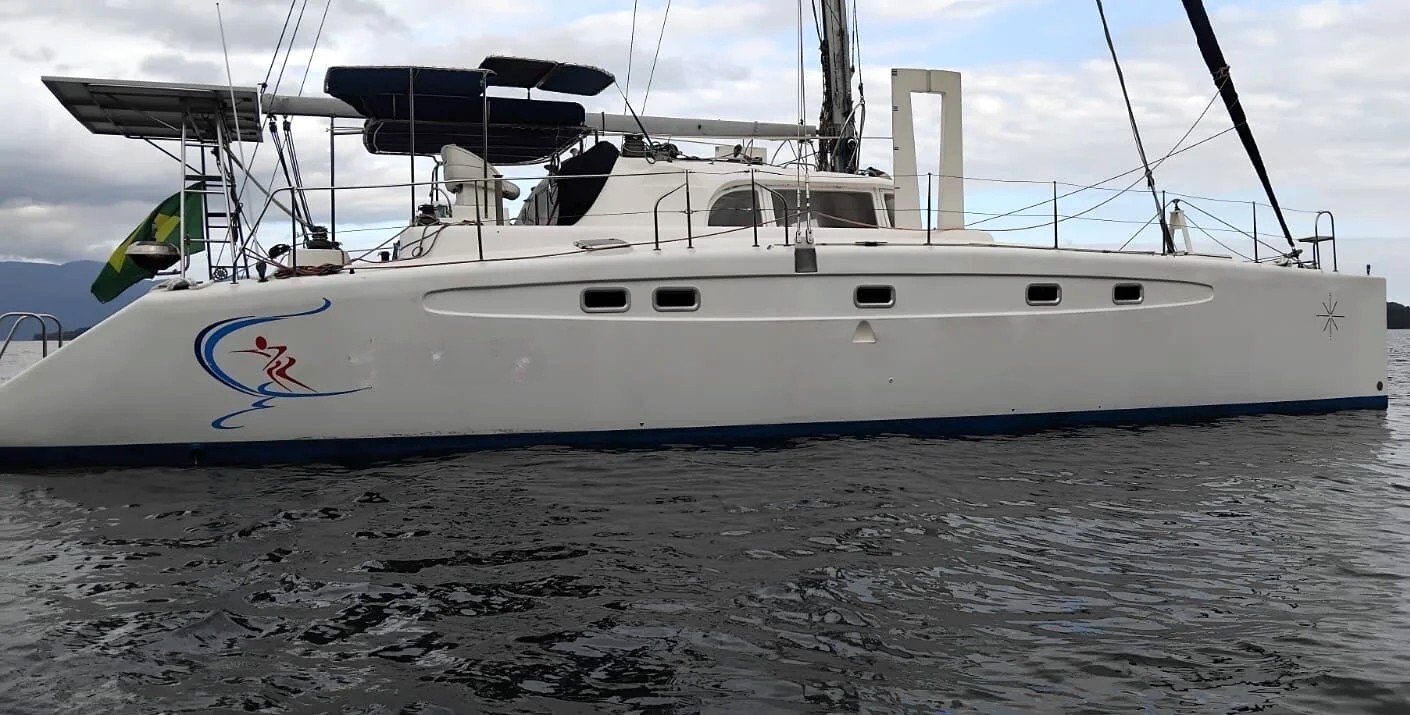 Catamarã Dolphin 46 fundeado, visto de perfil, com mastro, bimini, painéis solares e bandeira do Brasil
