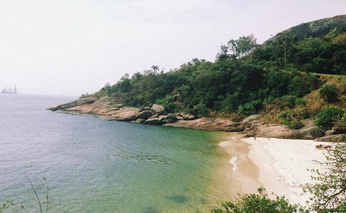 Praia Adão e Eva em Niterói, com água verde-esmeralda, areia branca e vegetação de Mata Atlântica ao fundo