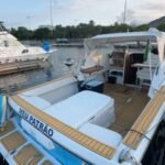 Vista de popa da lancha atracada na Marina da Glória, mostrando o cockpit, solário acolchoado, toldo e deck em teca, com o Rio de Janeiro ao fundo.