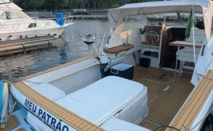Vista de popa da lancha atracada na Marina da Glória, mostrando o cockpit, solário acolchoado, toldo e deck em teca, com o Rio de Janeiro ao fundo.
