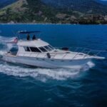 lancha Mares 45 navegando em mar azul, vista lateral com flybridge, montanhas e marina ao fundo